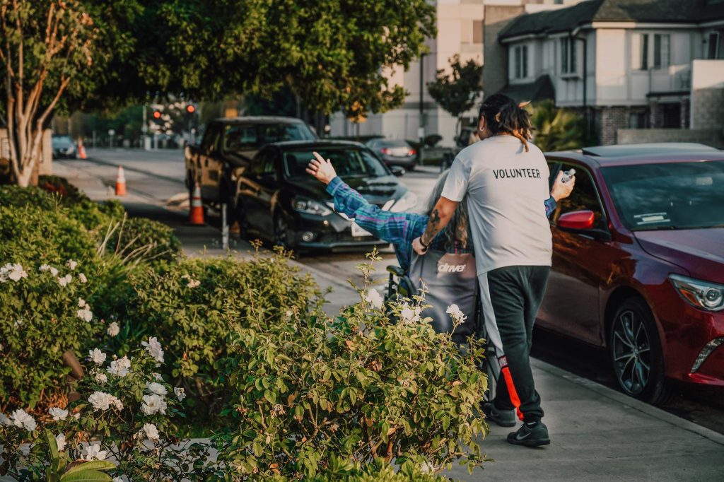 Volunteer assisting a person in a wheelchair on a city street surrounded by greenery and parked cars.
